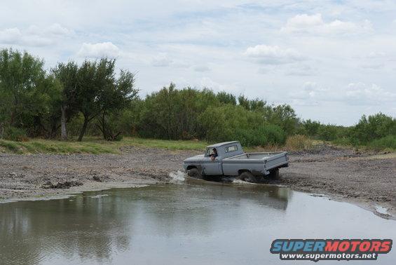 Gray pickup truck driving through a muddy puddle with trees in the background