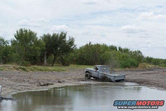 Gray pickup truck driving through a muddy puddle with trees in the background