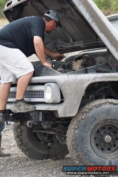 Gray 1970s Ford truck with hood open, man inspecting engine, mud on body and tires