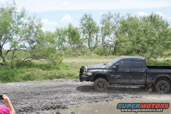 Black pickup truck with a brush guard driving through a muddy field with trees in the background