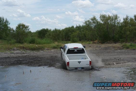 White pickup truck driving through a muddy puddle with trees in the background. Sticker on rear window reads 'WEEKEND 4 WHEELERS'