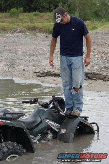 Dark green Can-Am Outlander ATV partially submerged in muddy water, with a man standing on its fender