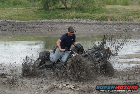 Man riding an ATV through a muddy puddle, kicking up a lot of spray