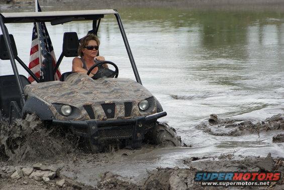 Woman driving a camouflage side-by-side UTV through mud, American flag visible