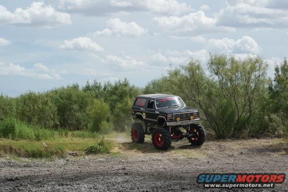 Monster truck with huge tires driving on a dirt path, trees and clouds in background. Has red rims