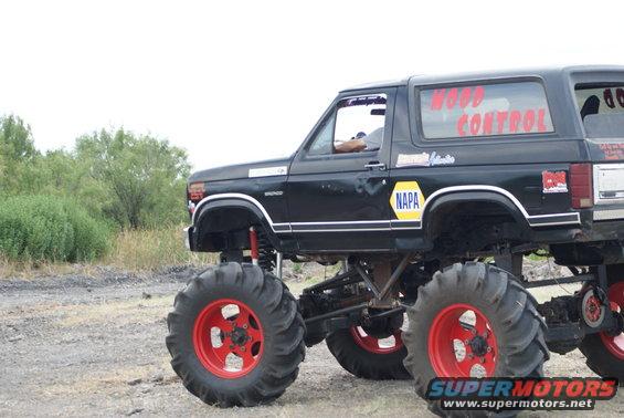 Black monster truck with red rims and large tires, lifted suspension, side view. "WOOD CONTROL" text on rear window