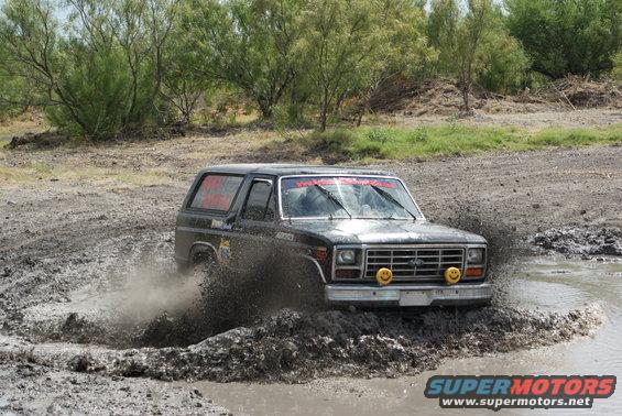 Black Ford Bronco splashing through a mud pit. Has smiley face lights on the front