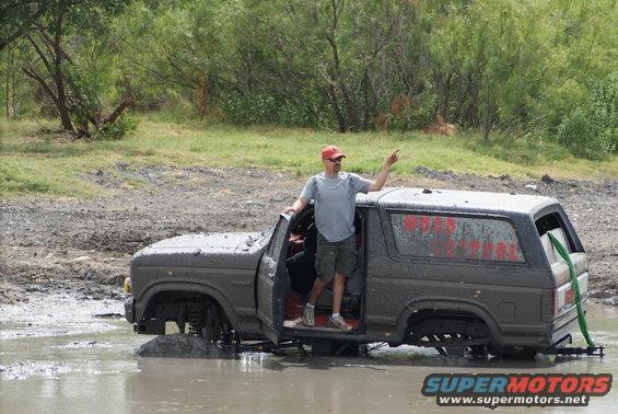 Man standing in a muddy 1989 Ford Bronco stuck in water, pointing. Red text on the side