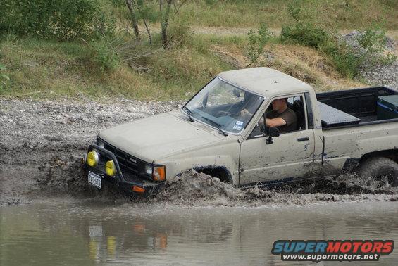 Tan Toyota pickup truck with yellow fog lights stuck in mud, driver visible. License plate reads TXT-575