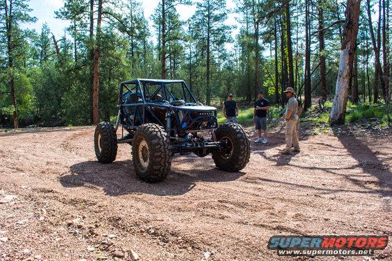 Custom buggy with roll cage and large tires parked on dirt road, trees in background, three people standing nearby