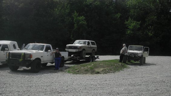White Ford Bronco on a trailer pulled by a white pickup truck. Another white Jeep is parked nearby