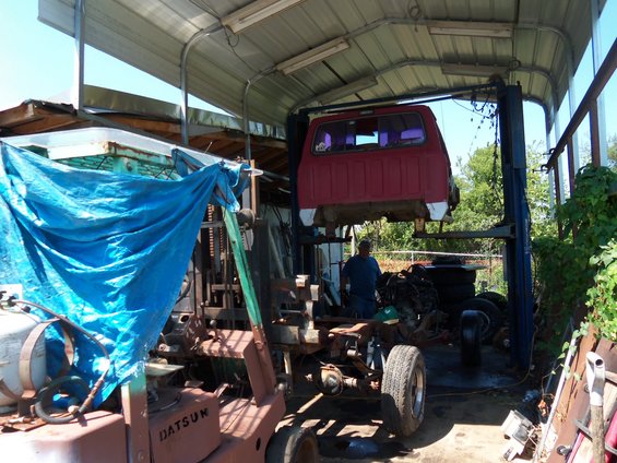 Red pickup truck cab suspended on a lift, with a forklift and disassembled vehicle parts in a workshop