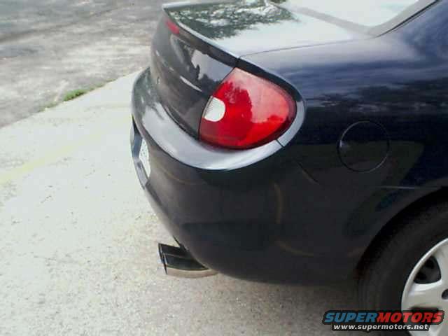 Dark sedan's rear quarter panel, taillight, and exhaust tip. The car appears to be a 2000 Dodge Neon