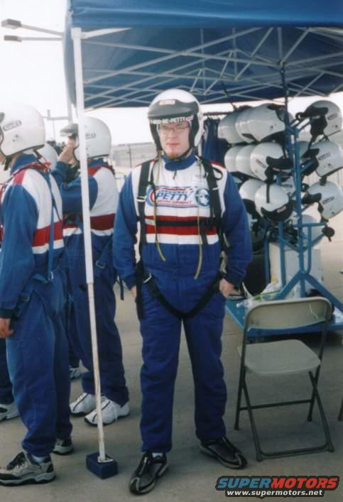 People in blue racing suits and helmets stand near a rack of helmets and a folding chair