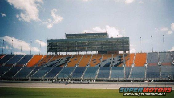 Racetrack grandstands with blue and orange seats under a cloudy sky. A large building with multiple levels is visible at the top