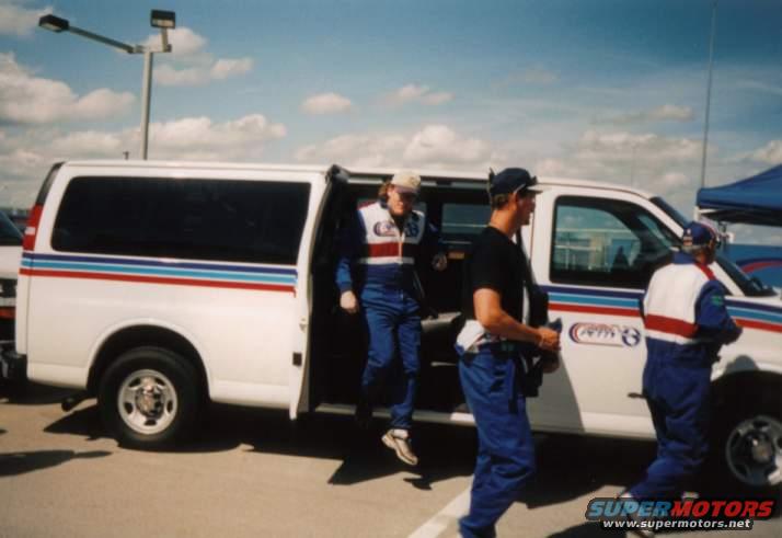 White passenger van with racing crew members getting out. One person in a racing suit is stepping out of the open side door