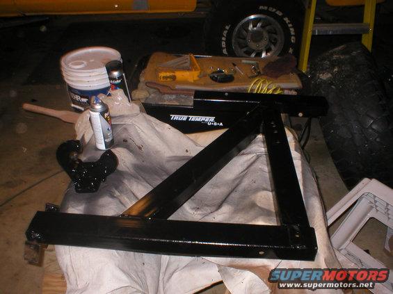 Black metal trailer hitch receiver and parts on a drop cloth, with a bucket, spray cans, and a toolbox nearby