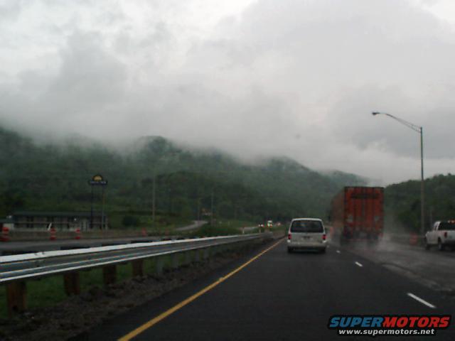 Highway with a white van, red truck, and white pickup truck under cloudy skies and misty mountains