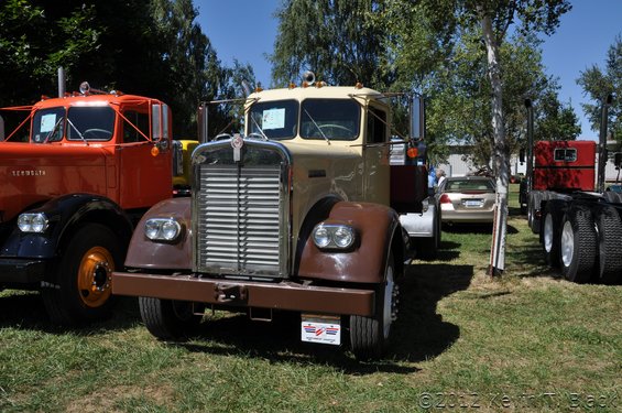 Front view of a tan Kenworth truck with a chrome grille, parked next to an orange truck and a red semi-truck. A car is visible behind it