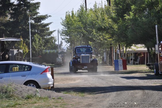 Front view of a dark-colored old truck driving on a dirt road, kicking up dust. A silver car is parked to the left