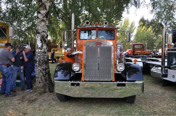Front view of an orange and black Peterbilt semi-truck with a large chrome bumper and grille. Other trucks and people are visible in the background