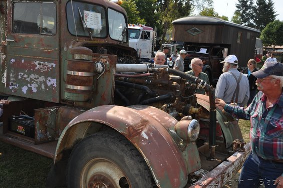 Rusted truck cab and exposed engine with people gathered around. Another truck and building are in the background