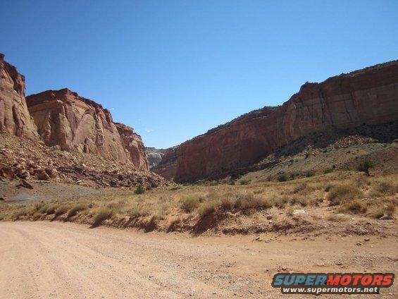 Dirt road leading into a canyon with red rock formations and dry brush under a clear blue sky