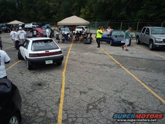 White hatchback with racing numbers on rear window, parked in a lot with other cars