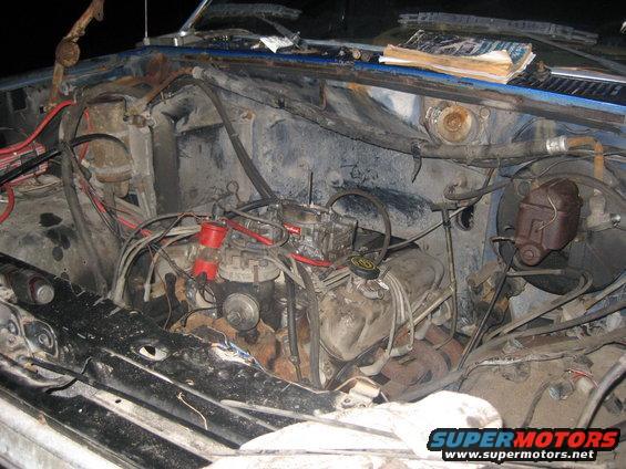 Engine bay of a blue vehicle with a V8 engine, carburetor, and visible wiring. The engine appears dirty and aged