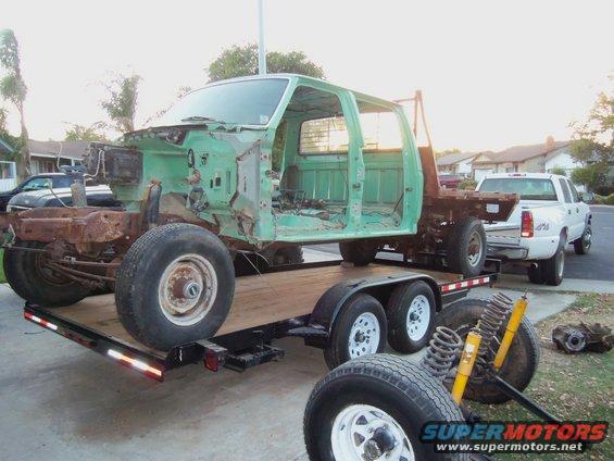 Green truck cab and chassis on a trailer, with a white pickup truck towing it. Loose suspension parts are on the ground
