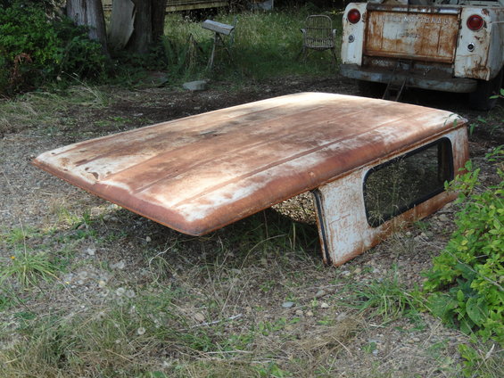 Rusted truck cap with a window, lying on the ground. A white pickup truck is visible in the background