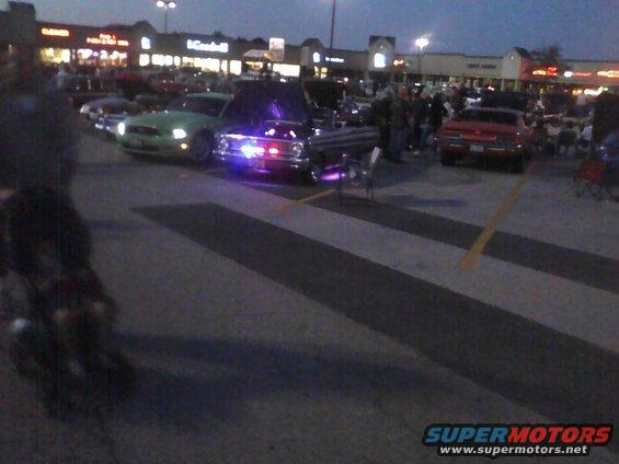 Cars gathered in a parking lot at dusk, some with hoods up and lights on