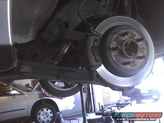 Rear brake rotor and caliper assembly on a vehicle lift. Another car is visible below