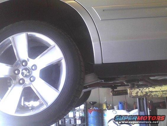 Close-up of a silver sedan's wheel and lower body on a lift. The wheel has five spokes and a center cap with a horse emblem