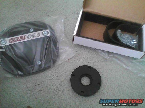 Two black circular parts, one in plastic packaging with 'PERFECT LAUNCH' text, and a small open box with a strap and screws
