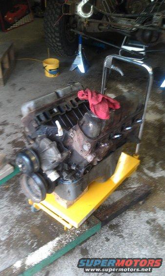 V8 engine on a yellow engine stand, red rag on top. Vehicle in background is on jack stands