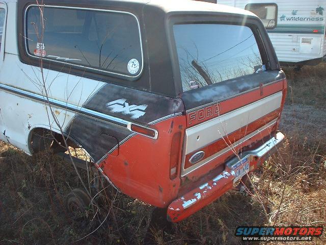 Rear view of a 1978 Ford Bronco with 4x4 graphics and a red, white, and black stripe. A trailer is parked behind it