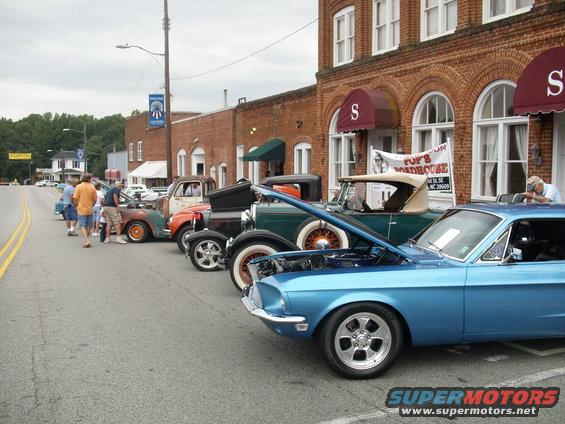 Several classic cars, including a blue Mustang with hood open, parked on a street in front of brick buildings