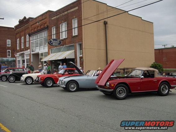 Several classic convertibles parked on a street in front of brick buildings. One red car has its hood open