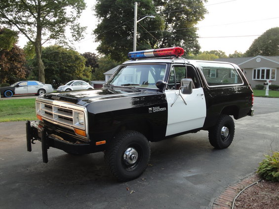 Black and white police SUV with light bar on top. Ramcharger badge visible. Other cars in background
