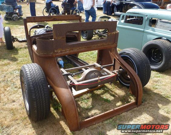 Rusted car chassis, rear view, with other vehicles in background
