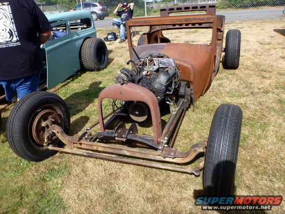 Rusty hot rod chassis with engine and front suspension visible. Another car is parked to the left