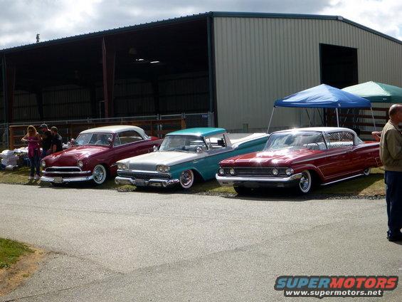 Three classic cars parked outside a building. The car on the right is a red 1959 Mercury Montclair
