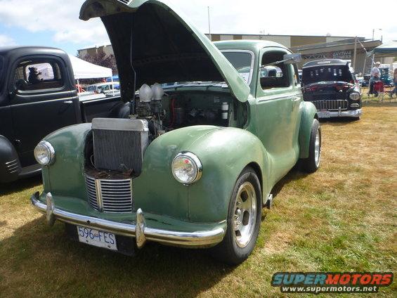 Green hot rod with hood open, showing engine. Another truck is to the left, and a dark car is behind. License plate reads 596-FES