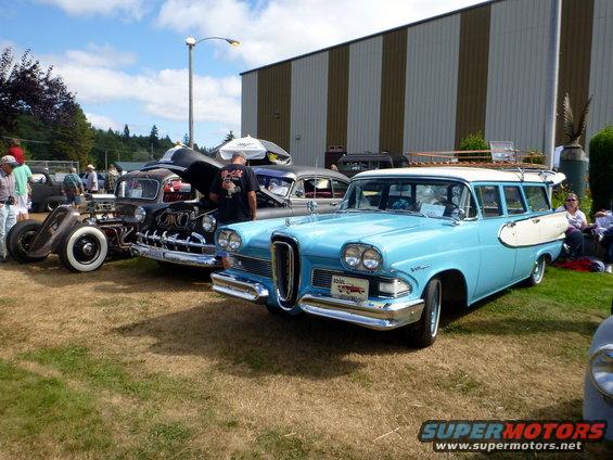 Blue and white station wagon with surfboard on the side, parked on grass with other cars