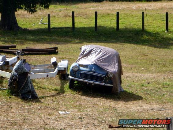 Blue car covered with a tarp parked next to a trailer in a grassy field