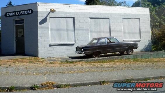 dark 1960s sedan parked next to a building with 'CAIN CUSTOM' sign