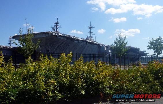 Aircraft carrier behind a fence and bushes under a blue sky with clouds