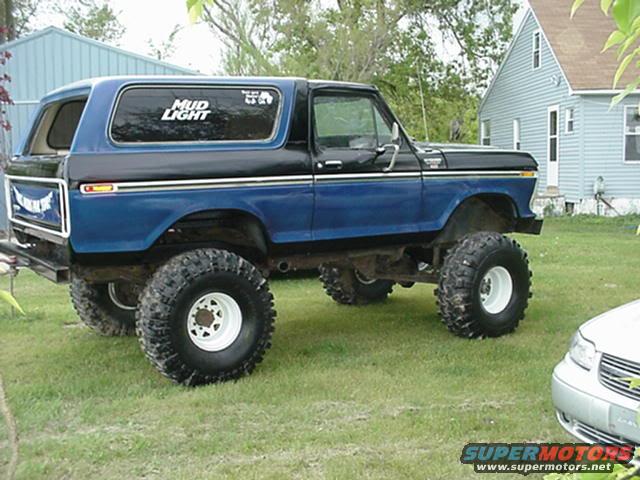 Blue and black lifted Ford Bronco with large tires, parked on grass in front of a house