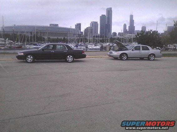 Two sedans parked by a harbor with city skyline in background. One has hood open